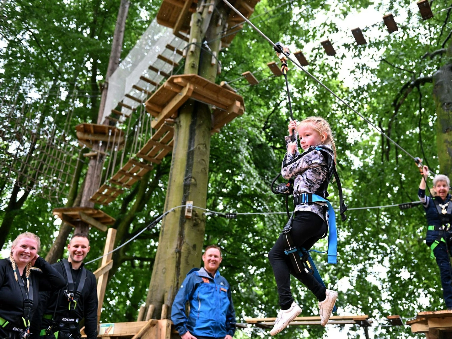 A young girl rides a Go Ape zip wire whilst her supervising adult watches on from a platform behind her, and her parents and a Go Ape instructor cheer her on from the ground.